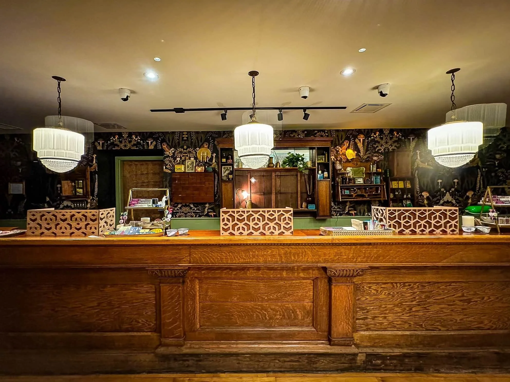 Interior view of a stylish reception area featuring an elegant wooden counter, decorative pendant lighting, and a vibrant floral wallpaper backdrop. The counter showcases patterned dividers and a display of brochures, creating a welcoming atmosphere for guests.