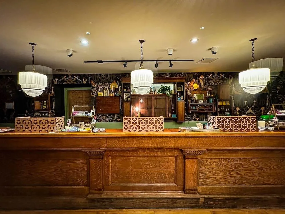 Interior view of a stylish reception area featuring an elegant wooden counter, decorative light fixtures, and a vibrant floral wallpaper backdrop, showcasing a welcoming atmosphere for guests.