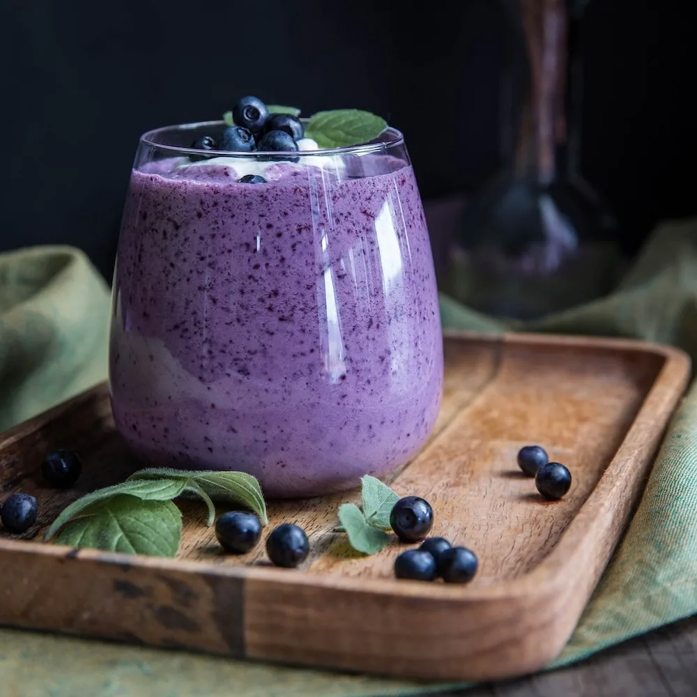 Creamy blueberry smoothie in a clear glass, garnished with fresh blueberries and mint leaves, served on a wooden tray with scattered blueberries and a green cloth background.