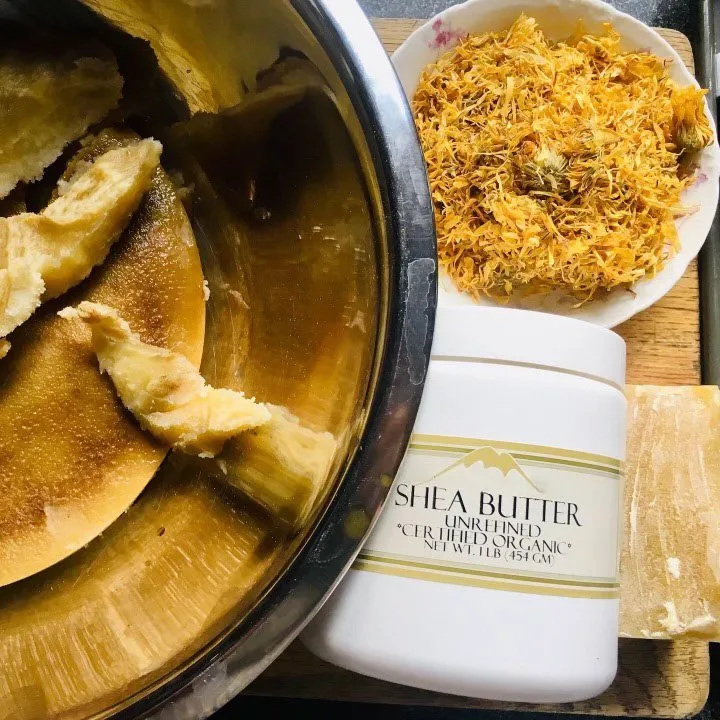 Bowl of unrefined shea butter next to a container labeled "Shea Butter Unrefined Certified Organic" and dried calendula flowers on a plate, showcasing natural skincare ingredients for homemade beauty products.