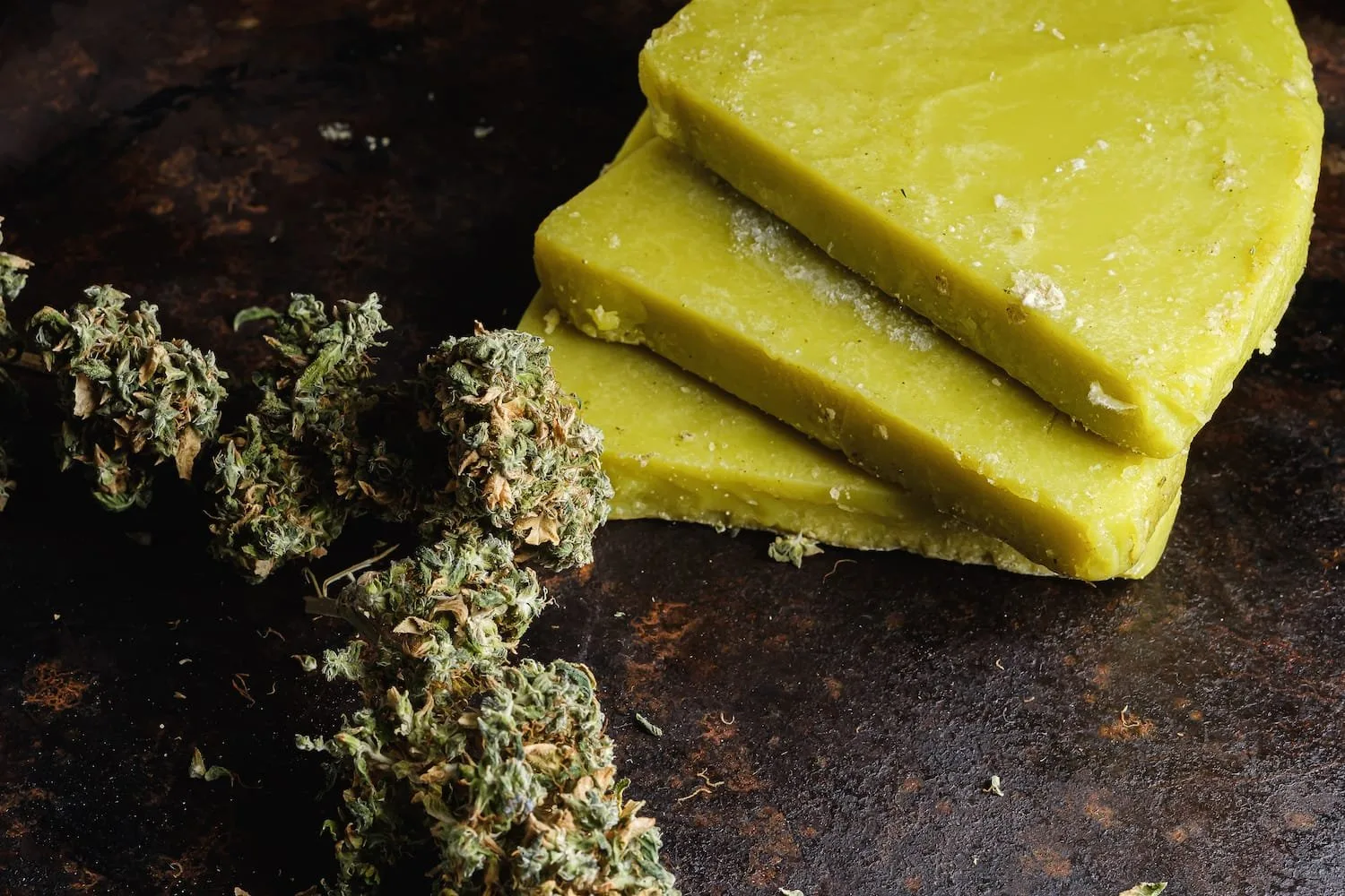 Close-up of three green cannabis wax slabs stacked on a dark surface, accompanied by dried cannabis buds, showcasing the texture and color of cannabis concentrates.