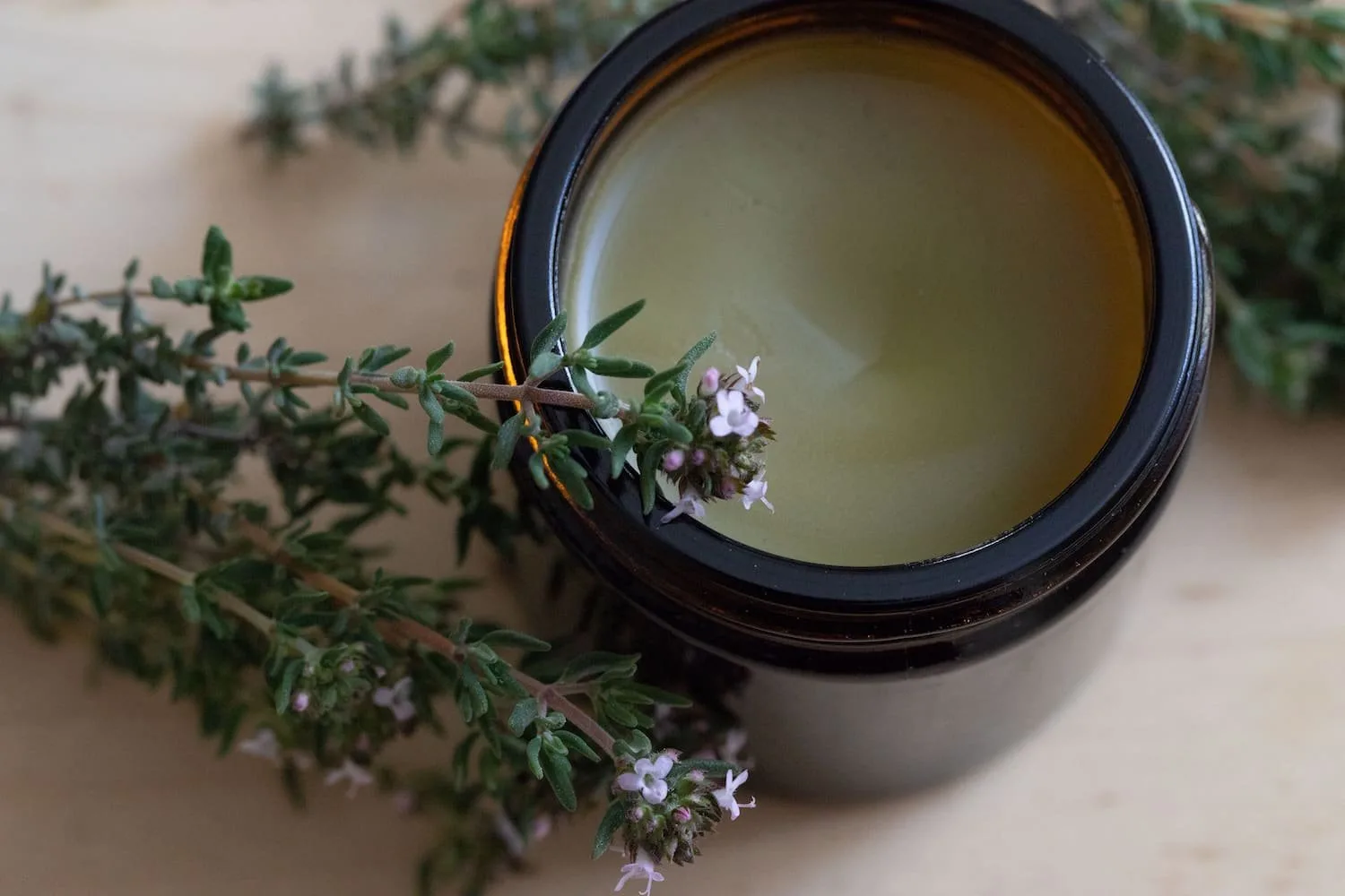 Amber glass jar filled with herbal balm, surrounded by fresh thyme sprigs and delicate purple flowers, set against a light wood background. Ideal for natural skincare and aromatherapy.