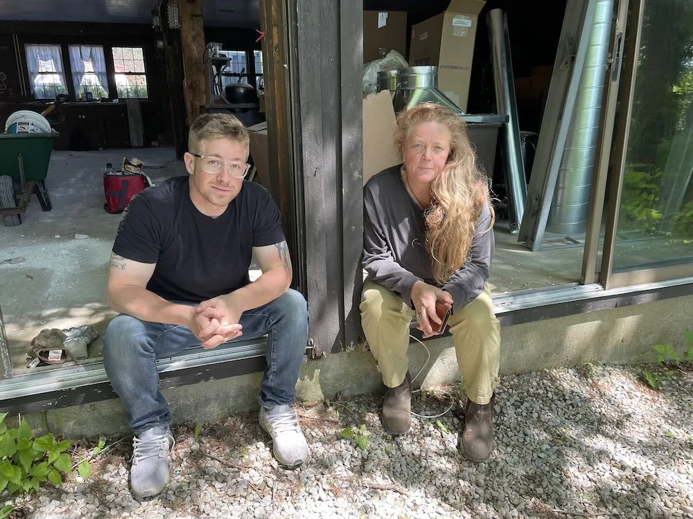 Two people sitting on the edge of a doorway outside a building under renovation, surrounded by tools and construction materials