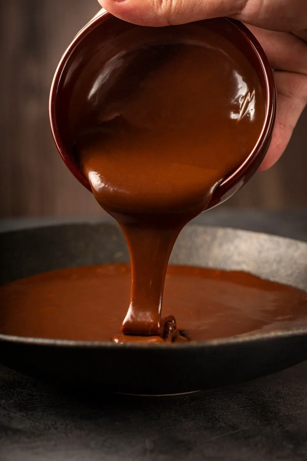 A hand pouring rich, glossy chocolate sauce from a bowl into a frying pan, showcasing the smooth texture and deep color of the sauce against a dark background.
