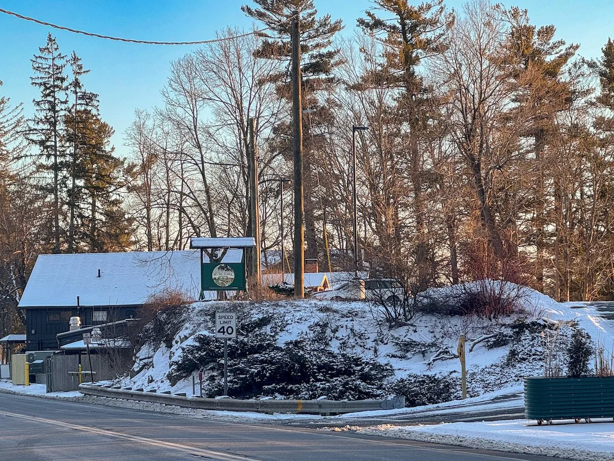 Snow-covered landscape featuring a quaint building and a green sign near a road with a speed limit of 40. Surrounding trees and a clear blue sky create a serene winter scene.