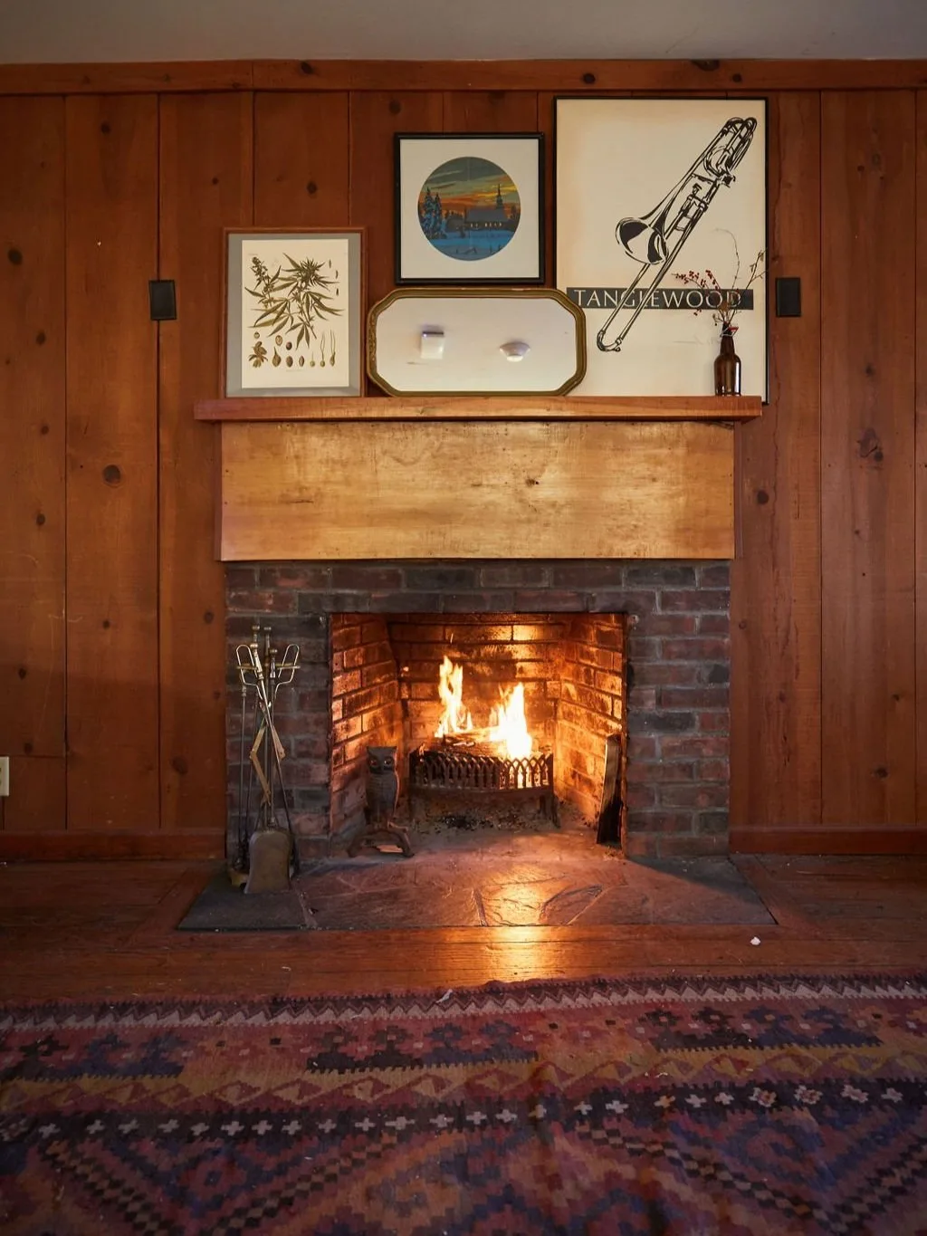 Cozy living room featuring a brick fireplace with a warm fire, surrounded by wooden paneling and a rustic mantel adorned with framed artwork and decorative items. A colorful area rug adds warmth to the hardwood floor.