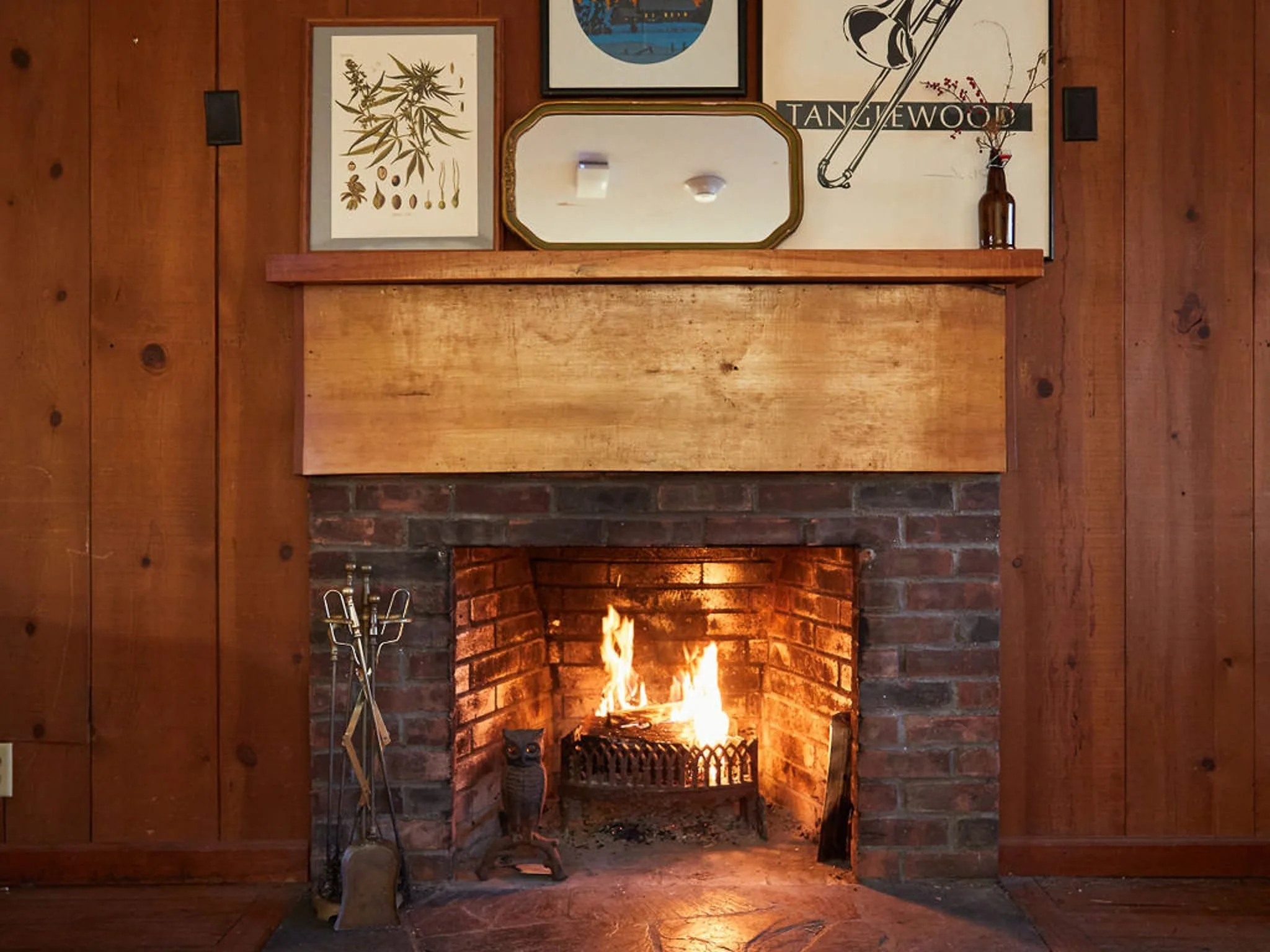 Cozy fireplace with a brick surround and wooden mantel, adorned with framed botanical art and a vintage poster. The warm glow of the fire adds a welcoming ambiance to the rustic wooden interior.