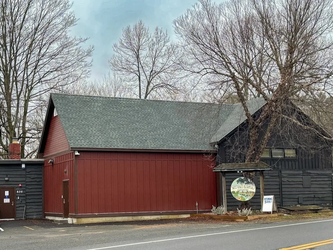 Exterior view of a rustic building featuring a red and black facade, surrounded by bare trees. A sign indicates the location is the "Short Road Botanical," with an "Opening Soon" notice displayed. The setting is along a quiet road, suggesting a peaceful, nature-oriented destination.