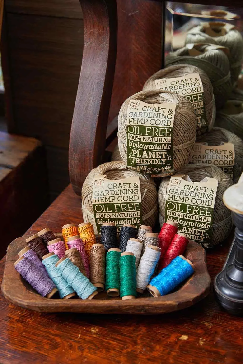 Colorful spools of thread in various shades arranged on a wooden tray, with balls of biodegradable hemp cord labeled as "Craft & Gardening Hemp Cord" in the background, emphasizing eco-friendly crafting supplies.