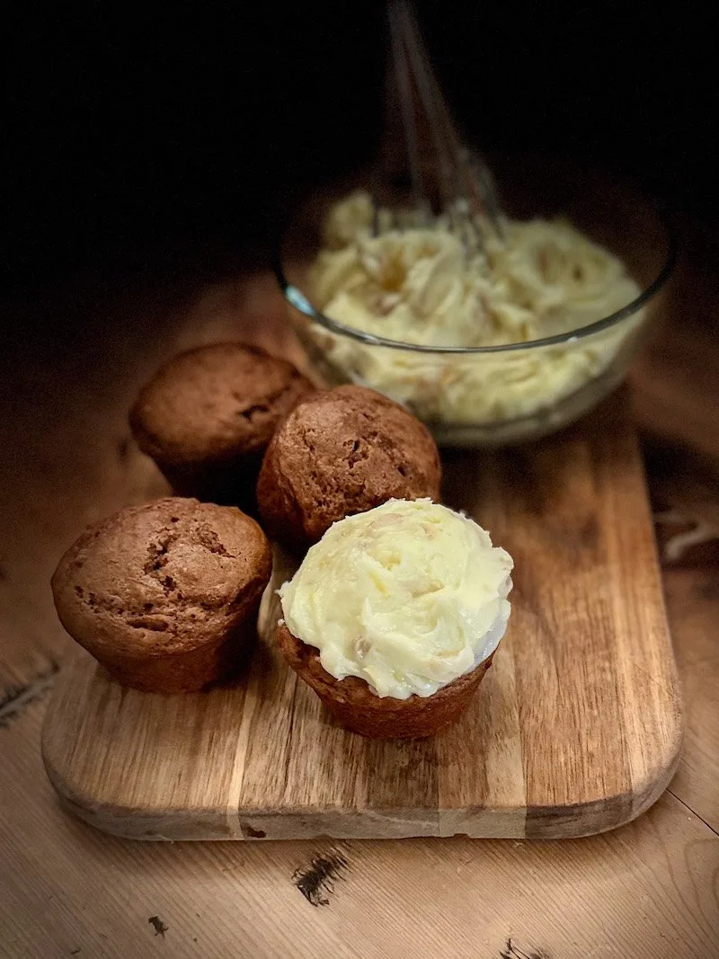 Freshly baked muffins topped with creamy buttercream frosting, arranged on a wooden cutting board, with a bowl of whipped frosting in the background.