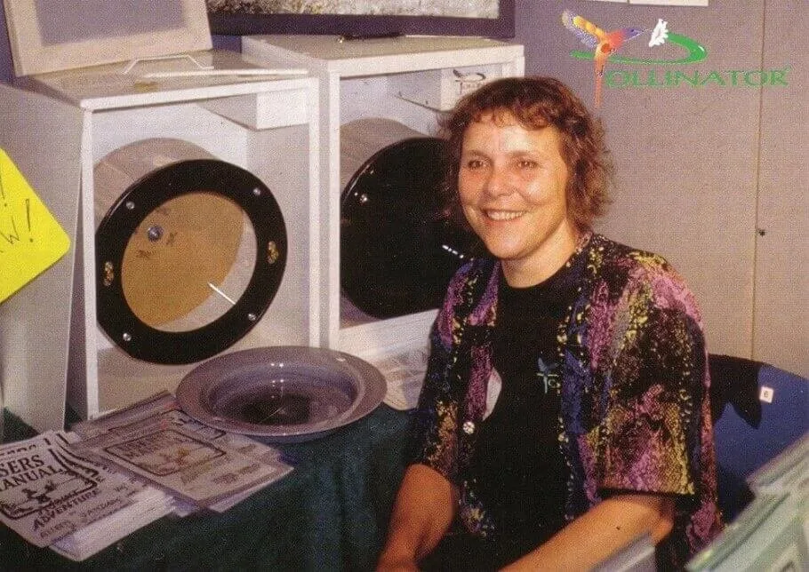 Smiling woman sitting at a booth with equipment displays and manuals, wearing a colorful patterned shirt