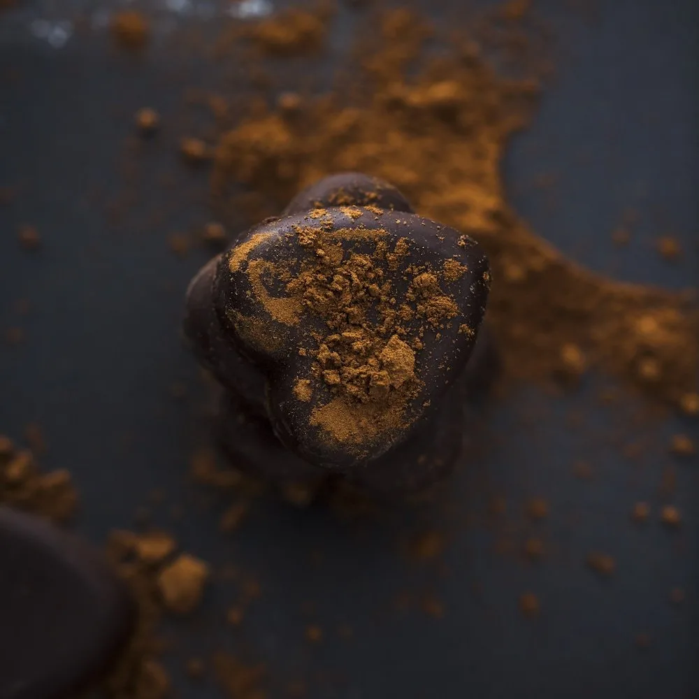 Dark chocolate heart-shaped treat dusted with golden spice powder, set against a dark background with scattered cocoa powder. Perfect for dessert recipes or gourmet chocolate lovers.