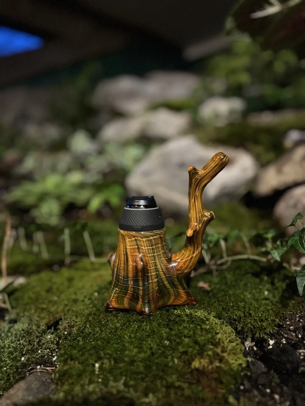 Unique glass bong designed to resemble a tree stump, featuring intricate wood-like patterns and a black bowl, set against a lush mossy background. Ideal for nature-themed smoking accessories.