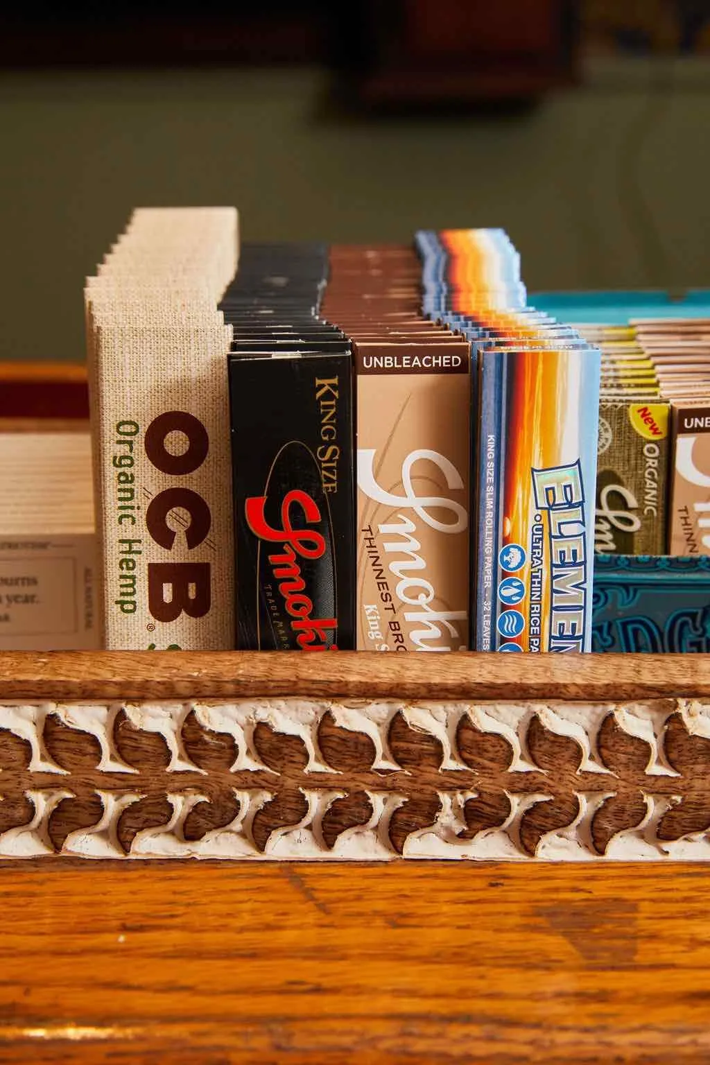 Display of various rolling papers including OCB Organic Hemp, Smoky King Size, and Elements ultra-thin rice papers, arranged neatly in a wooden holder with intricate carvings.