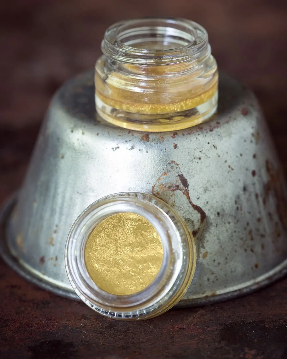 Glass jars containing a golden substance, placed on a rustic, weathered metal surface. The image highlights the texture and color of the contents, showcasing a creative or artisanal product.