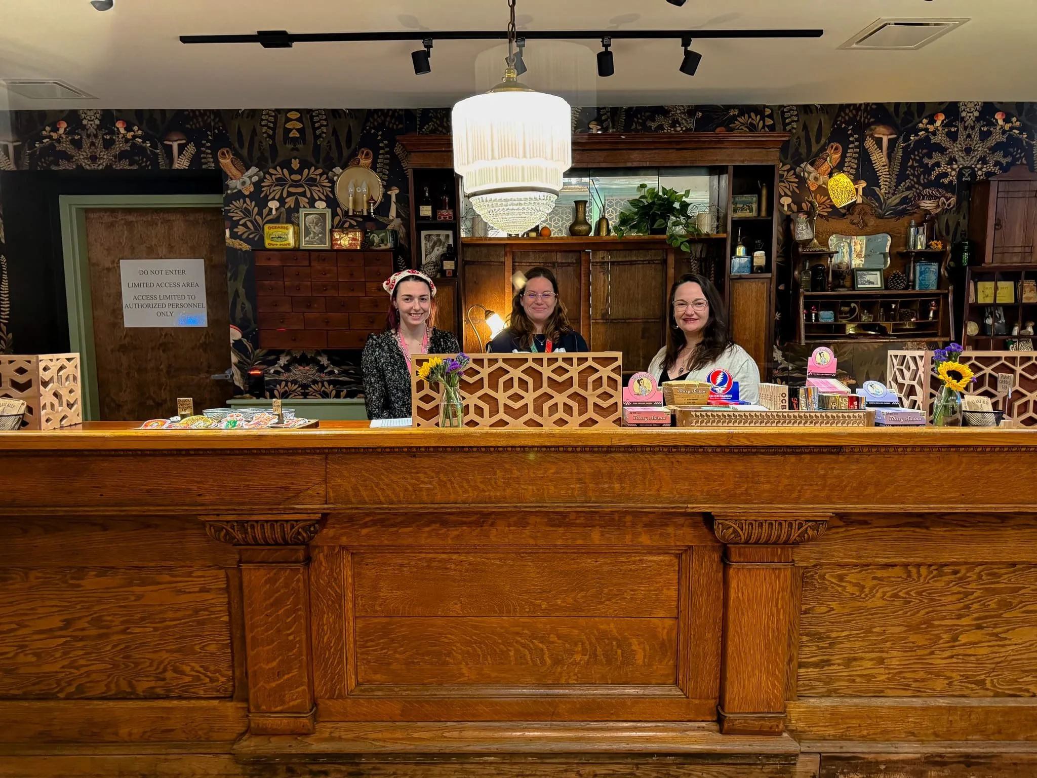 Three friendly staff members stand behind a wooden counter in a cozy, vintage-style shop adorned with floral wallpaper. The counter features colorful merchandise, including candies and decorative items, while a sign indicates a restricted access area. Sunflowers and other decorations enhance the inviting atmosphere.